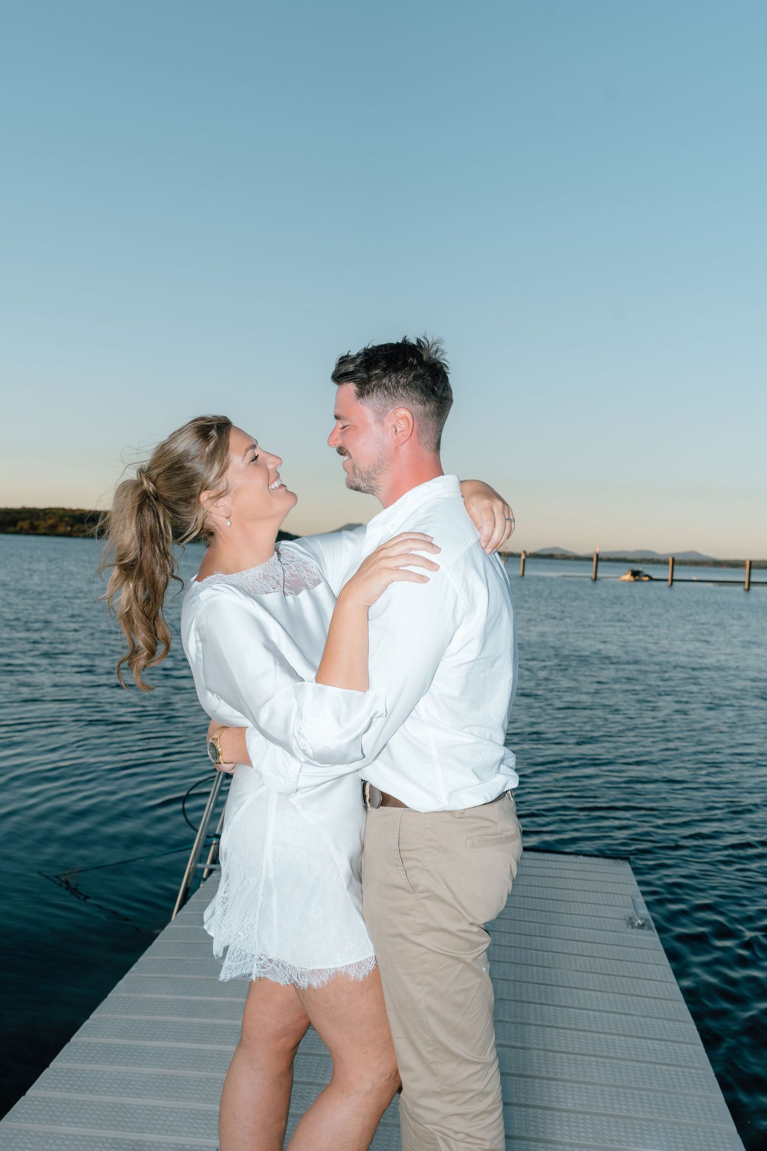 Bride and groom embrace and smile at each other on wooden jetty at golden hour sunset.