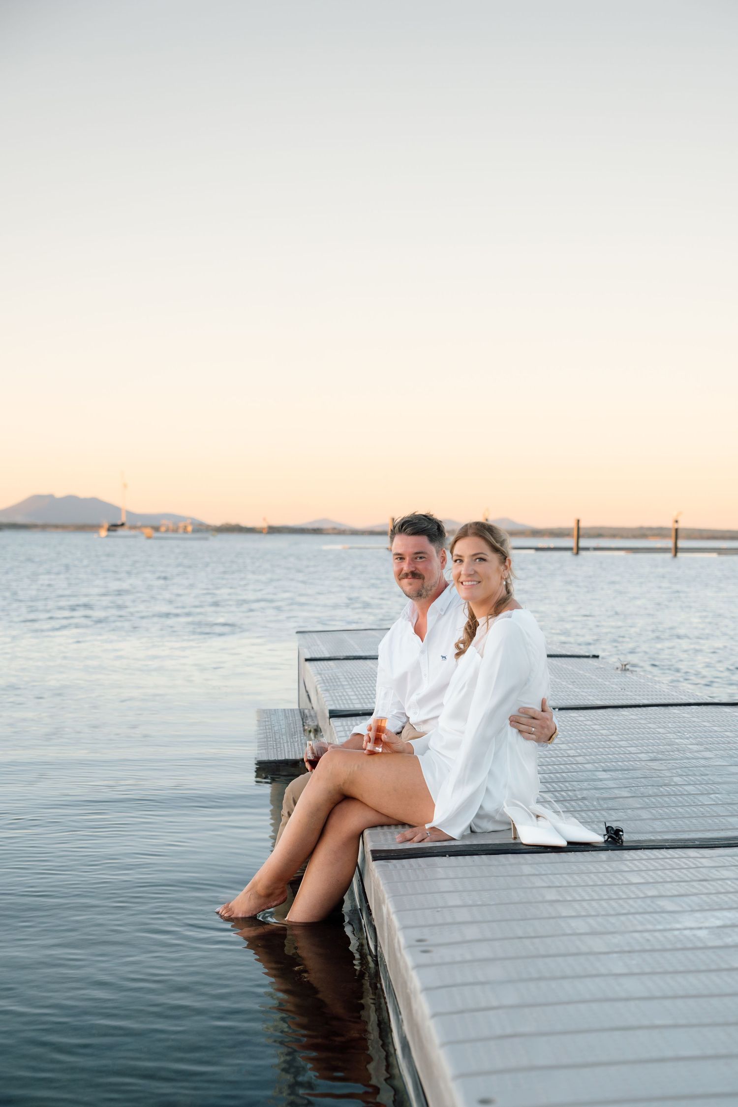 Couple in white robes sitting on a dock at sunset over calm water.