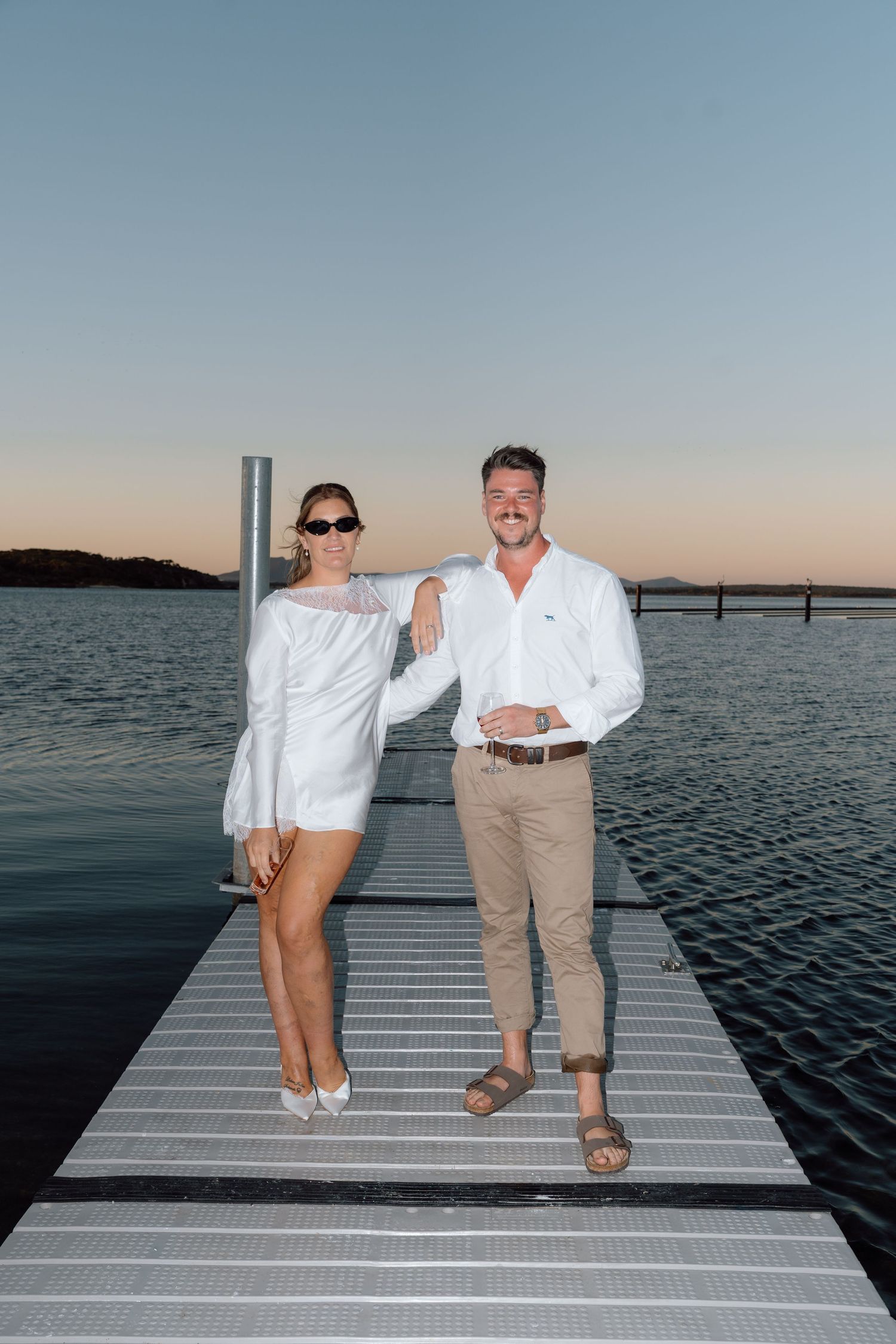Couple in white outfits posing on a dock at sunset over calm water.