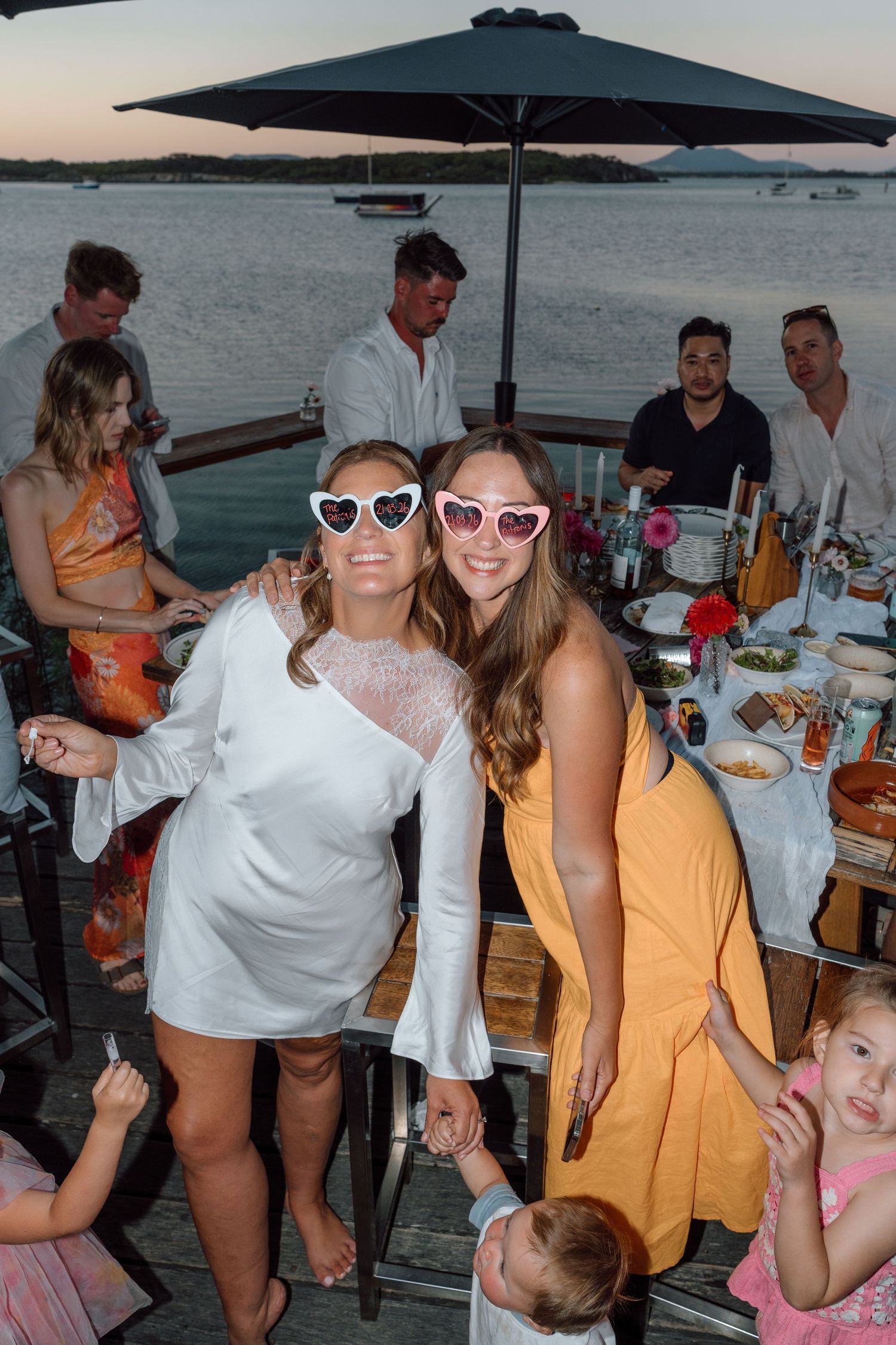 Two women in heart sunglasses smile at a waterfront dinner party at dusk.
