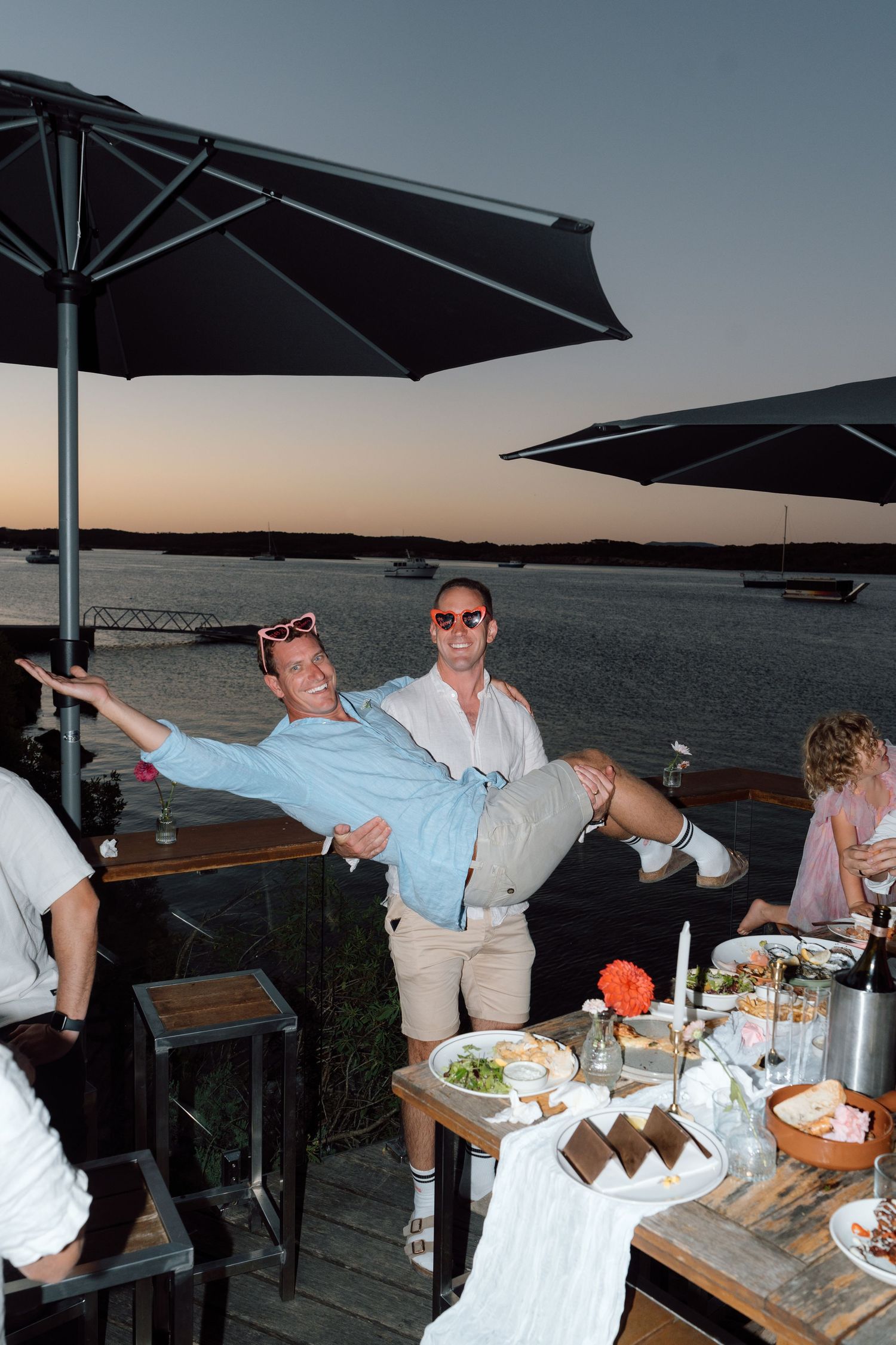 Two men joking at waterfront restaurant during sunset, one man being carried horizontally.