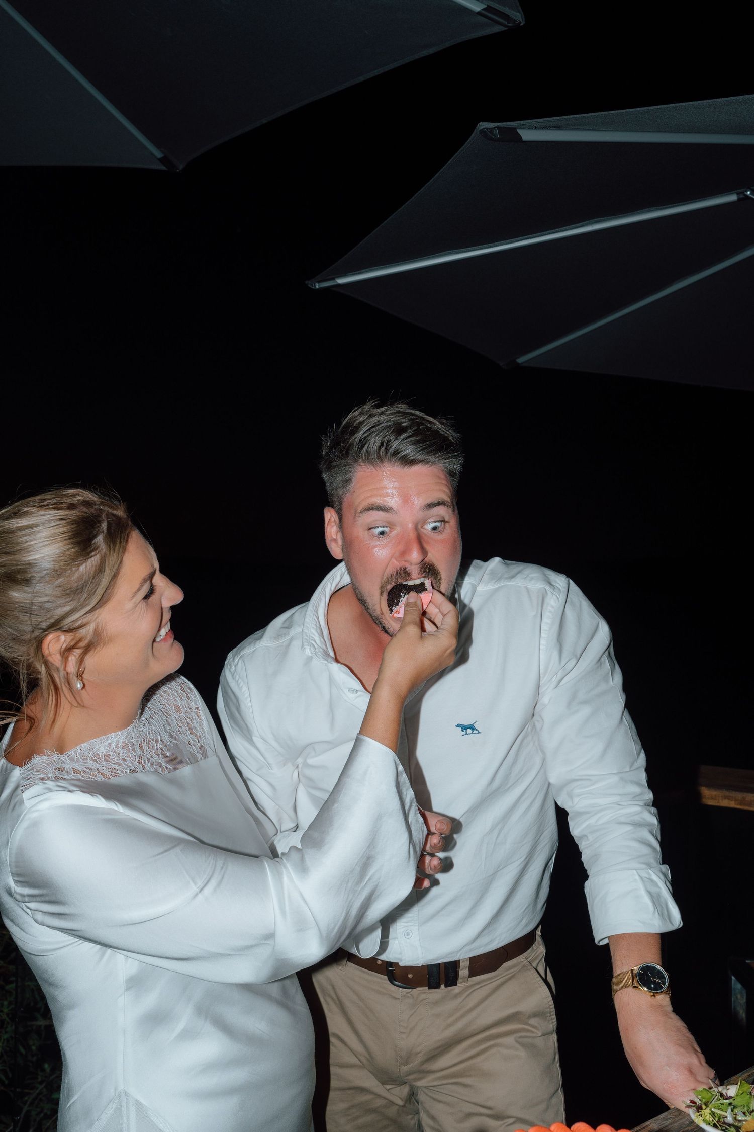 Bride feeds groom wedding cake at evening reception, both laughing joyfully in candid moment.