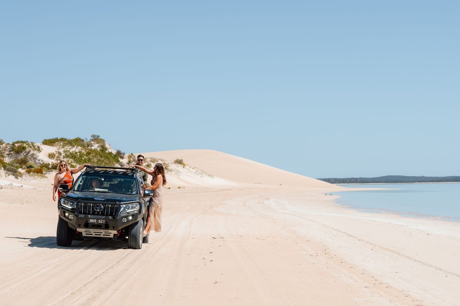 Group of friends posing beside a black 4WD truck on a white sandy beach with large sand dunes and blue water.