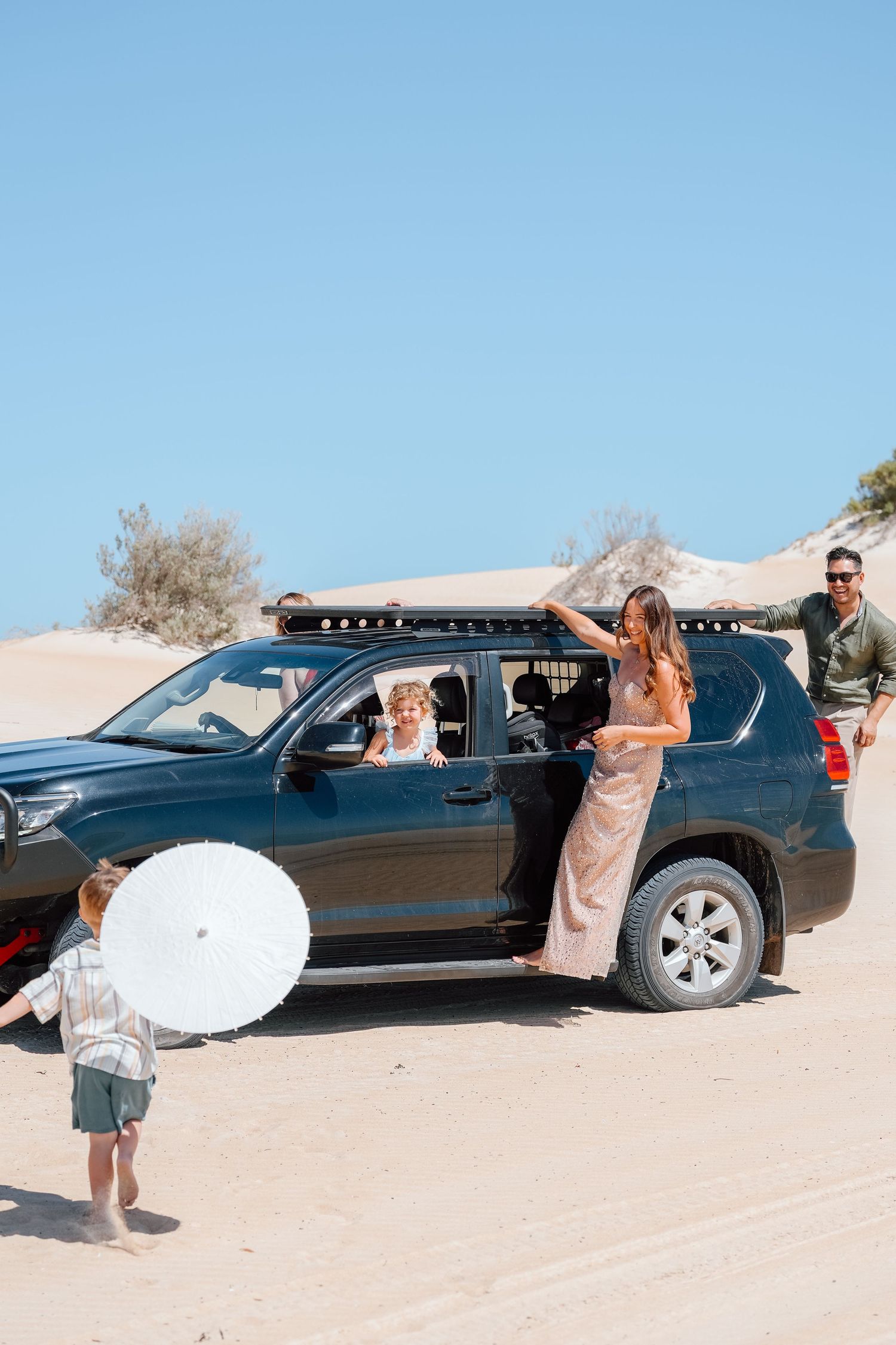 Family with children unloading from black 4WD SUV on sandy beach dunes on sunny day.