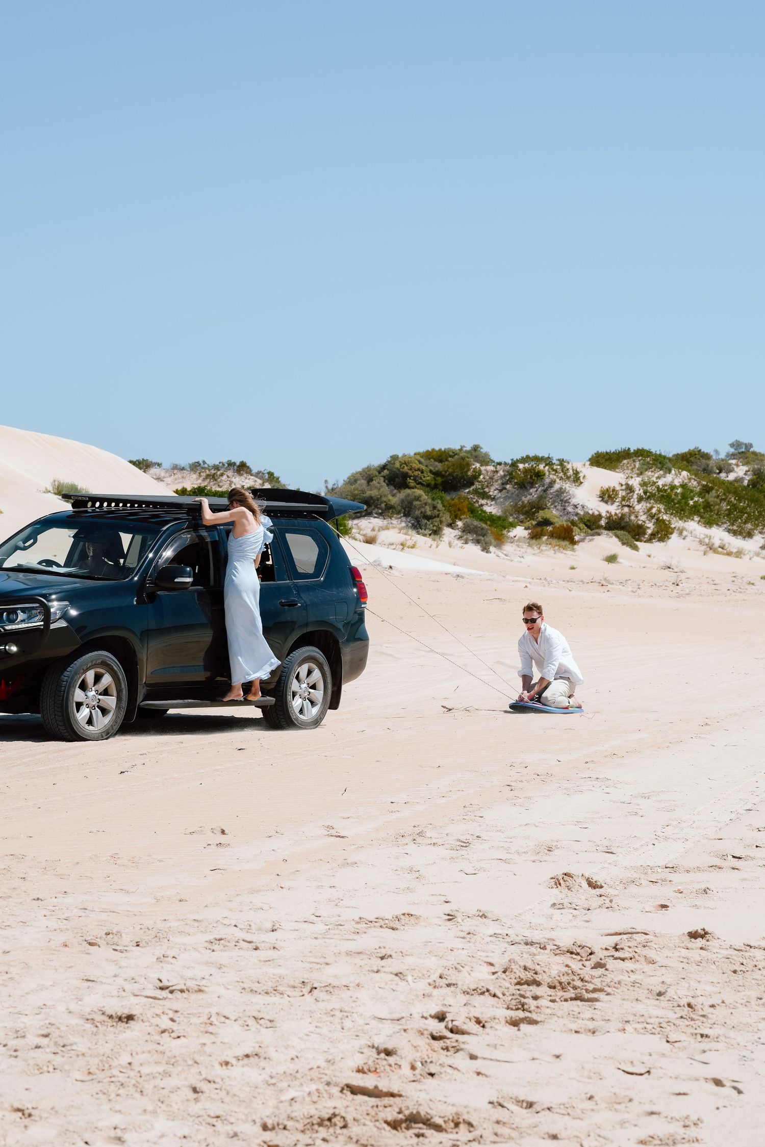 Couple in white outfits near black SUV on sandy dunes, woman leaning on car door, man crouching on beach.