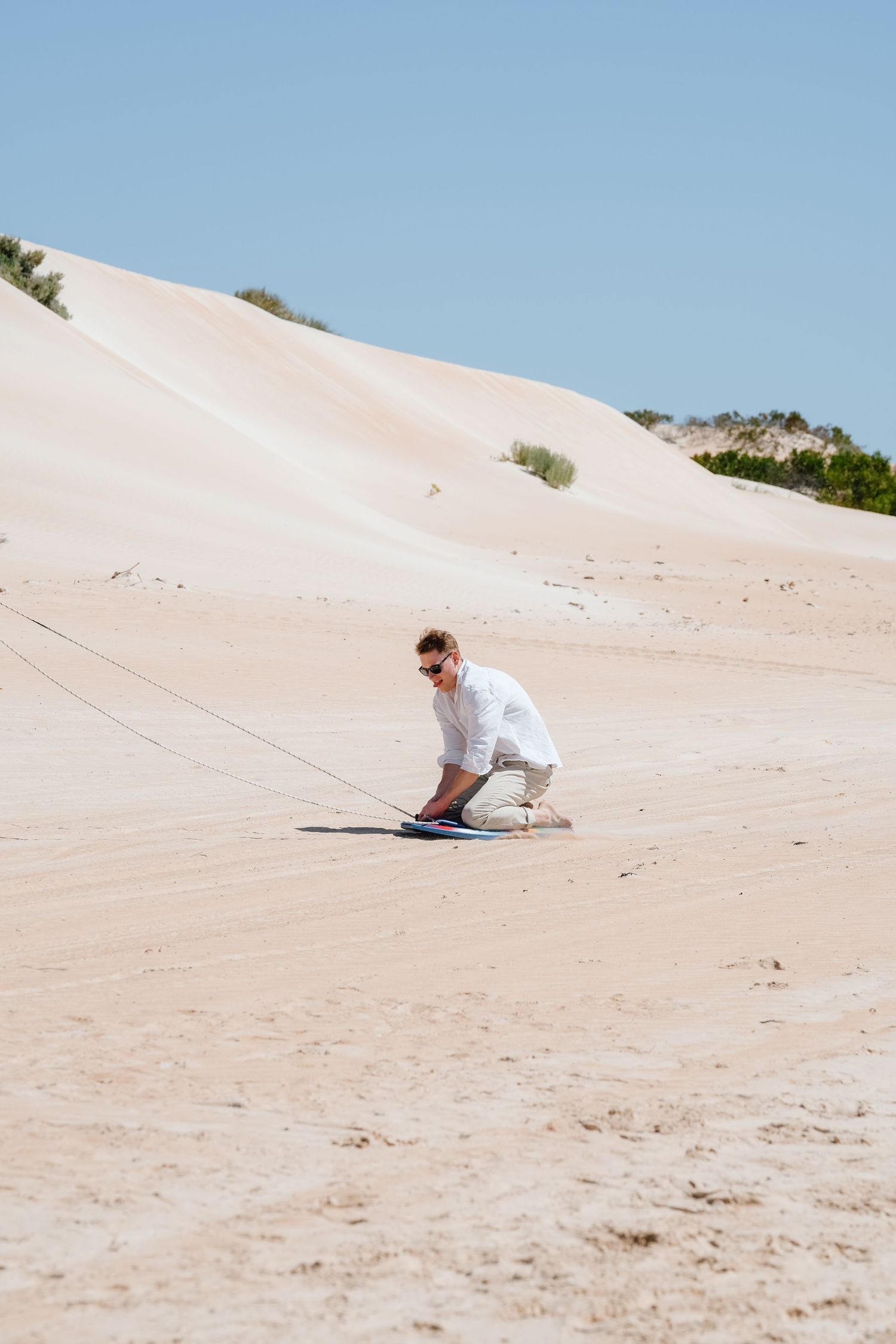 Man in white shirt sandboarding down a sandy desert dune on a sunny day with clear blue sky.
