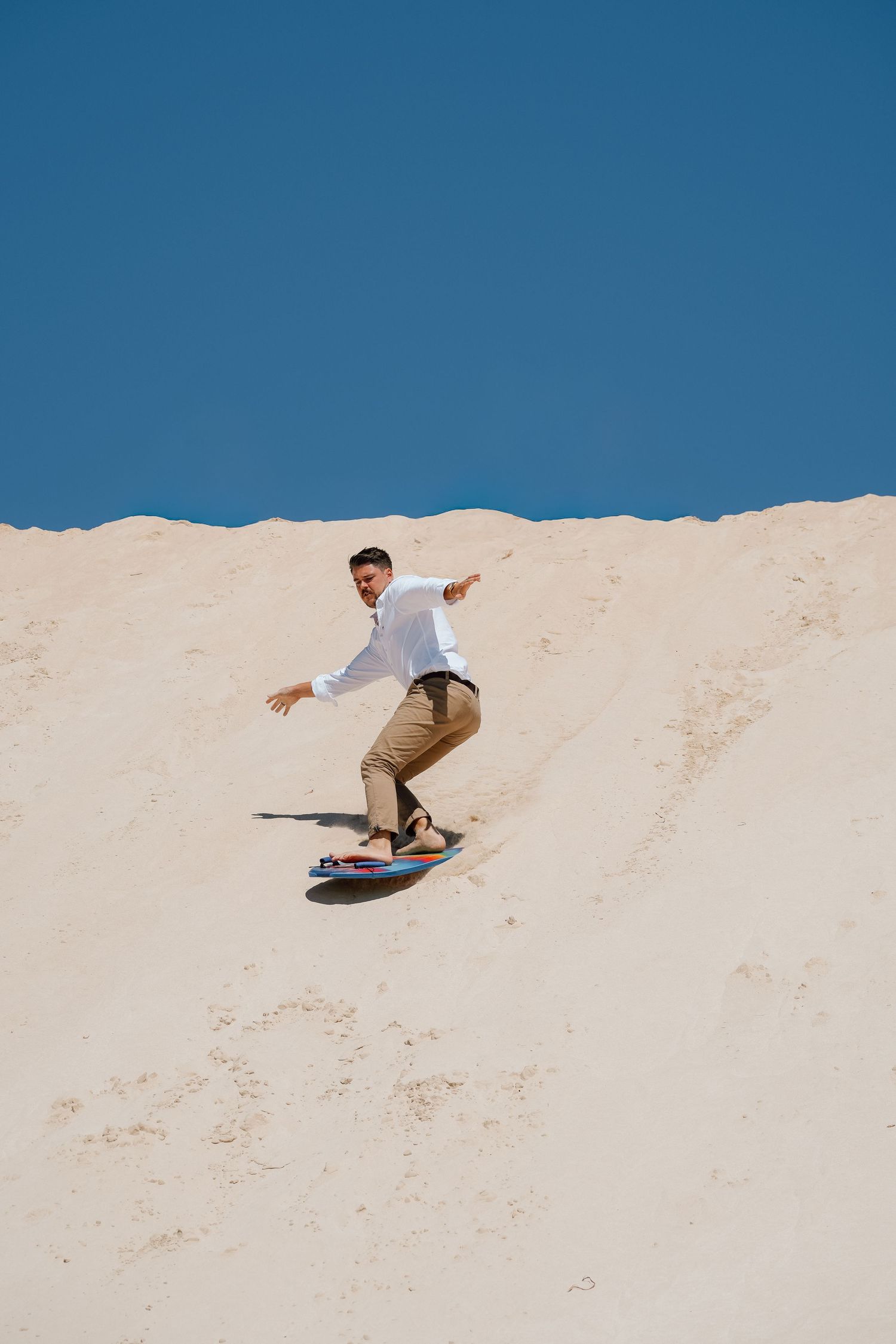 Man in white shirt sandboarding down a sandy dune under a clear blue sky.