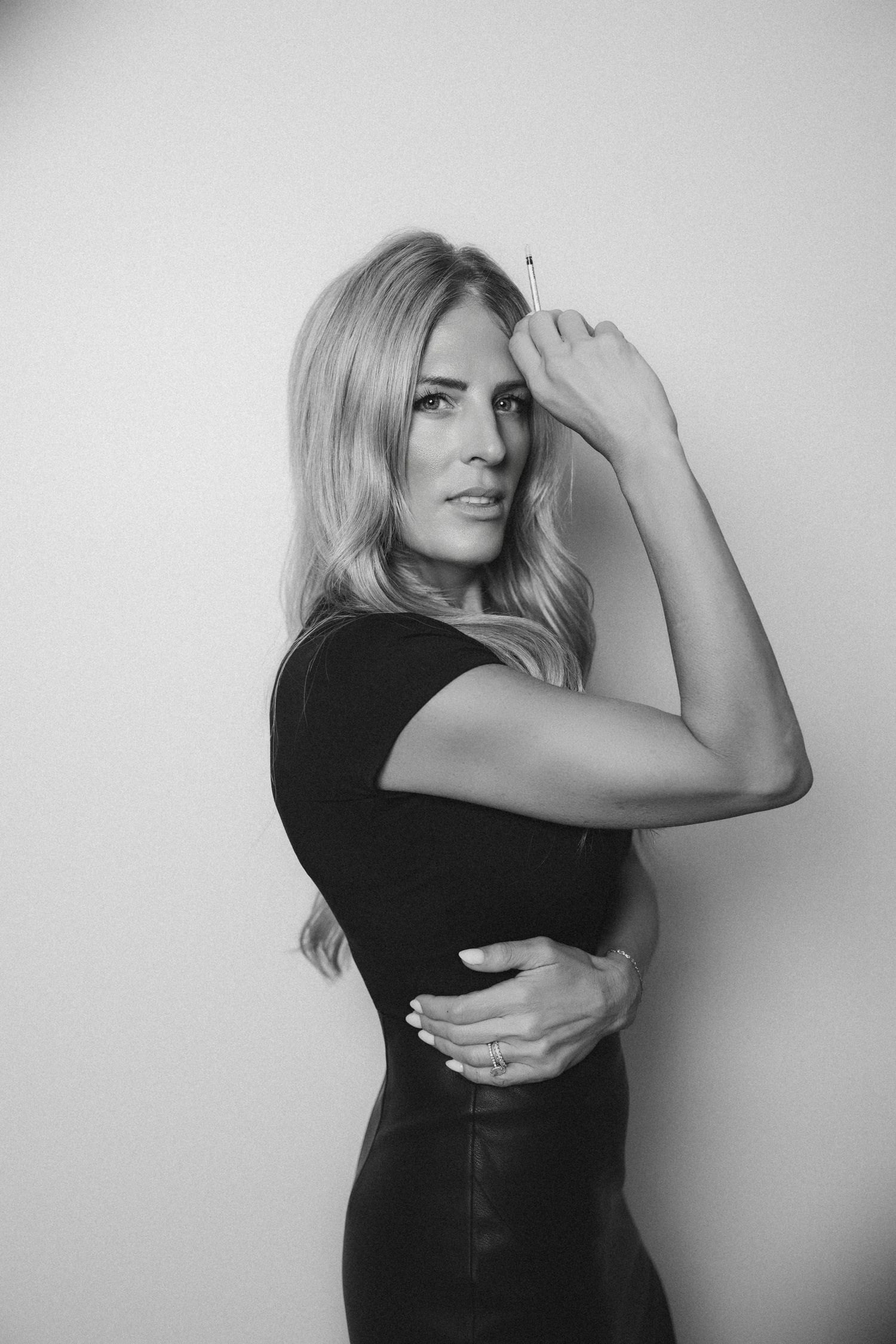 Black and white portrait of a woman with long wavy hair in a black dress, flexing her arm in a confident pose against a plain wall.
