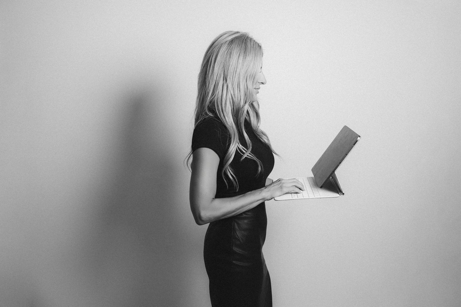 Black and white profile photo of a woman with long wavy hair in a fitted dark dress standing and reading a tablet against a plain wall.