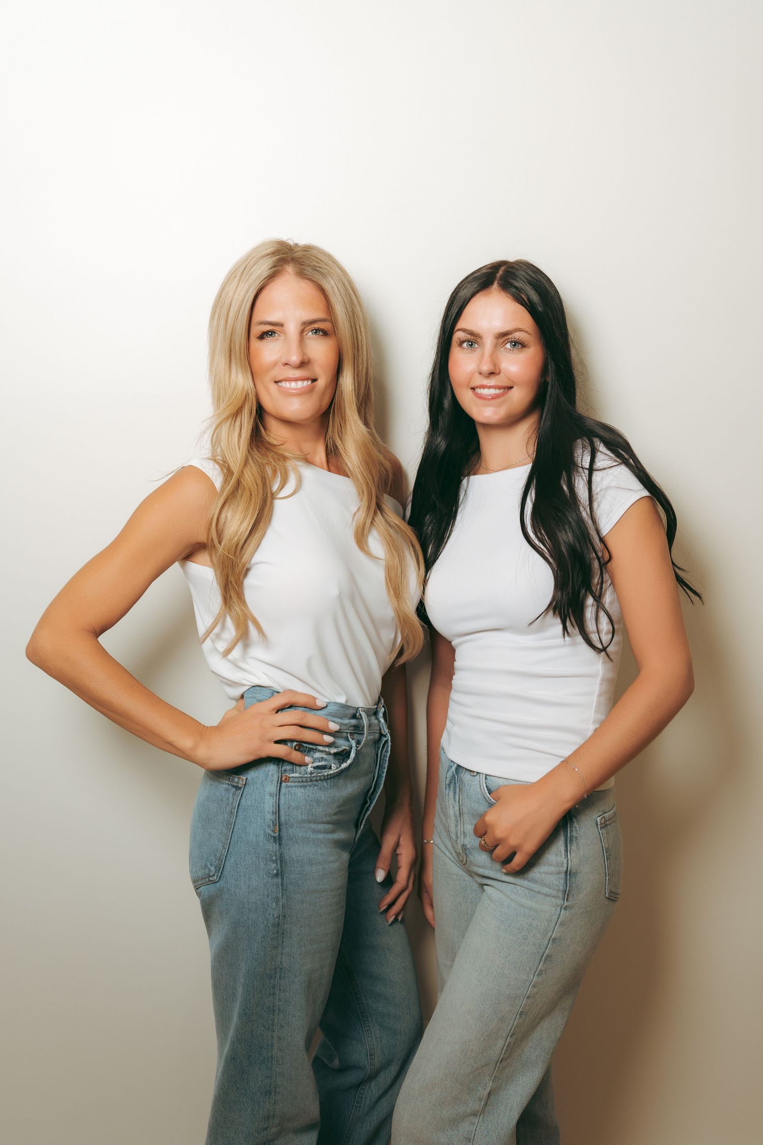 Two smiling women in white t-shirts and light blue jeans pose side by side against a neutral studio backdrop, one blonde and one brunette.