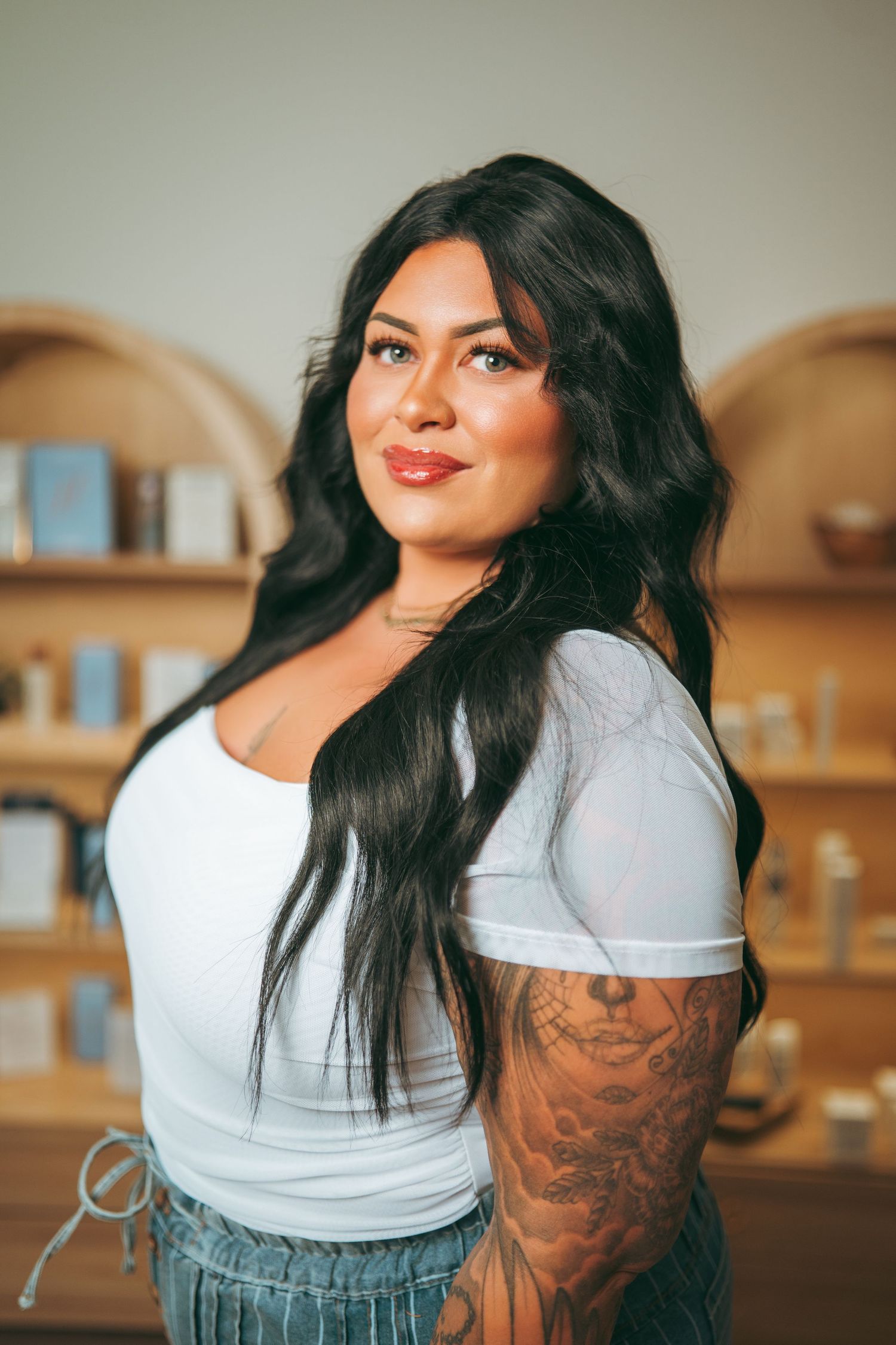 Woman with long wavy black hair, red lipstick and a floral arm tattoo smiling in a white top beside wooden shelves of skincare products.