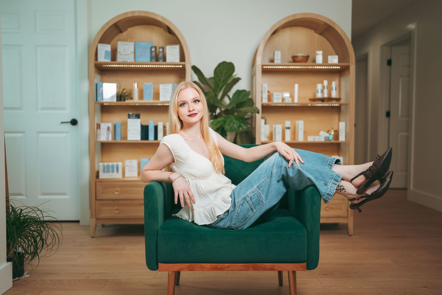 Woman with long blonde hair in a white top and jeans reclines on a green velvet armchair in front of wooden bookshelves and a potted plant.