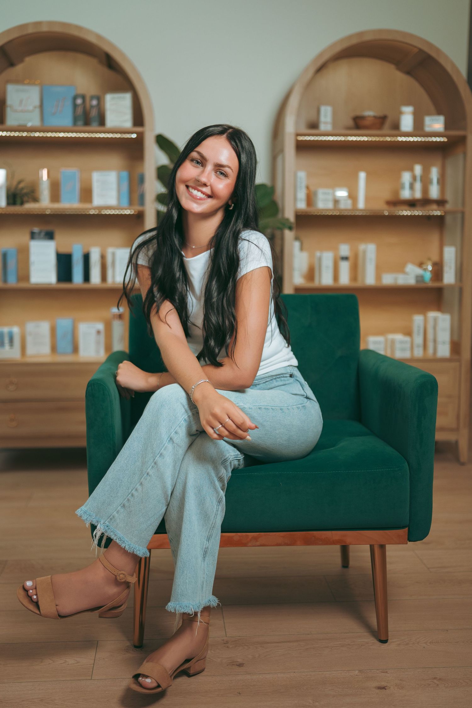 Smiling woman with long dark hair sits on a green velvet armchair in a boutique with wooden arched shelves of skincare products.