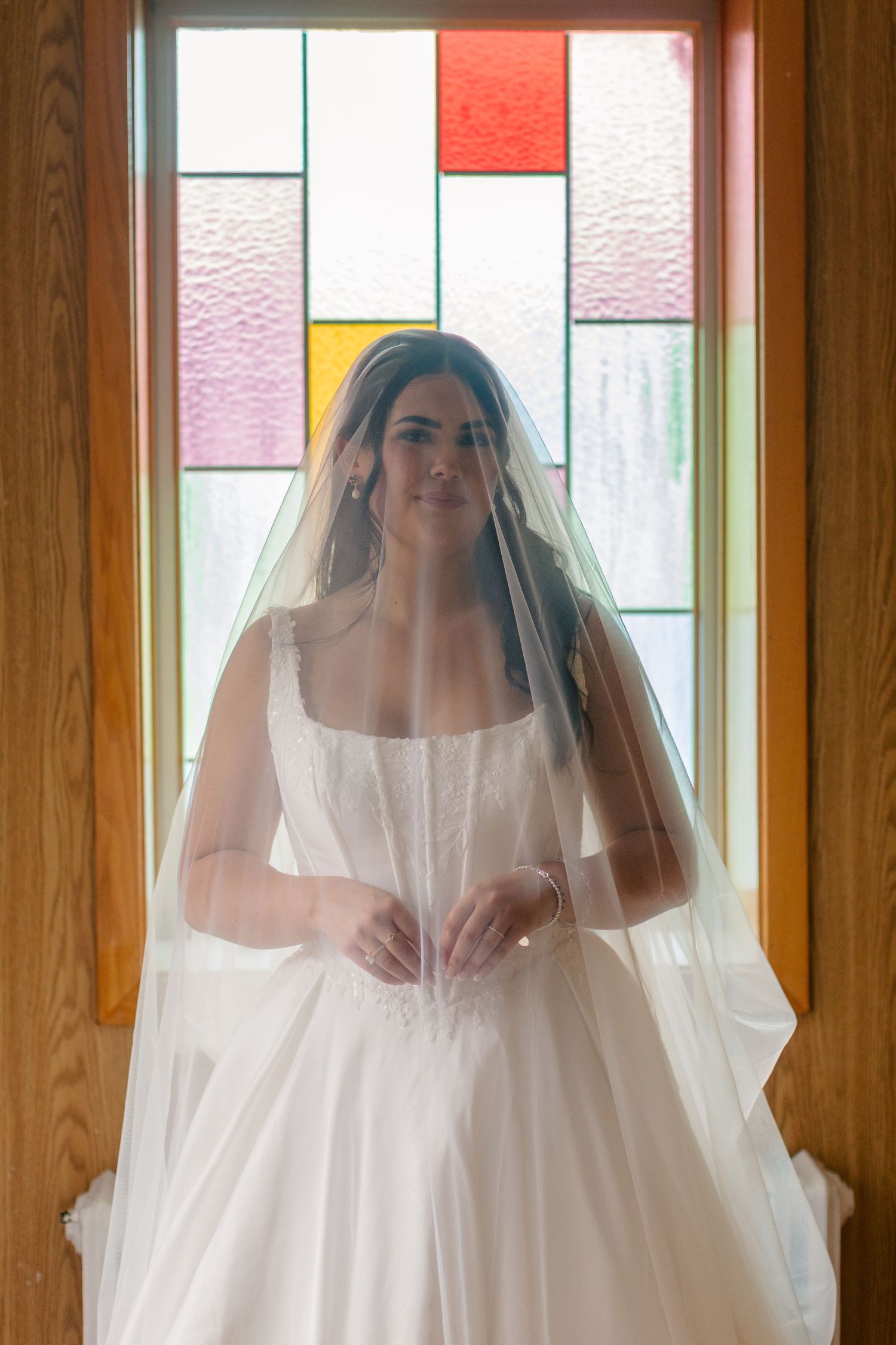 Bride in a white satin gown and sheer veil stands in front of a colorful stained glass window, holding her hands together at her waist.