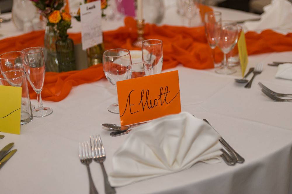 Elegant wedding reception table with orange Elliott place card, white napkin, silverware, and glassware on white tablecloth.