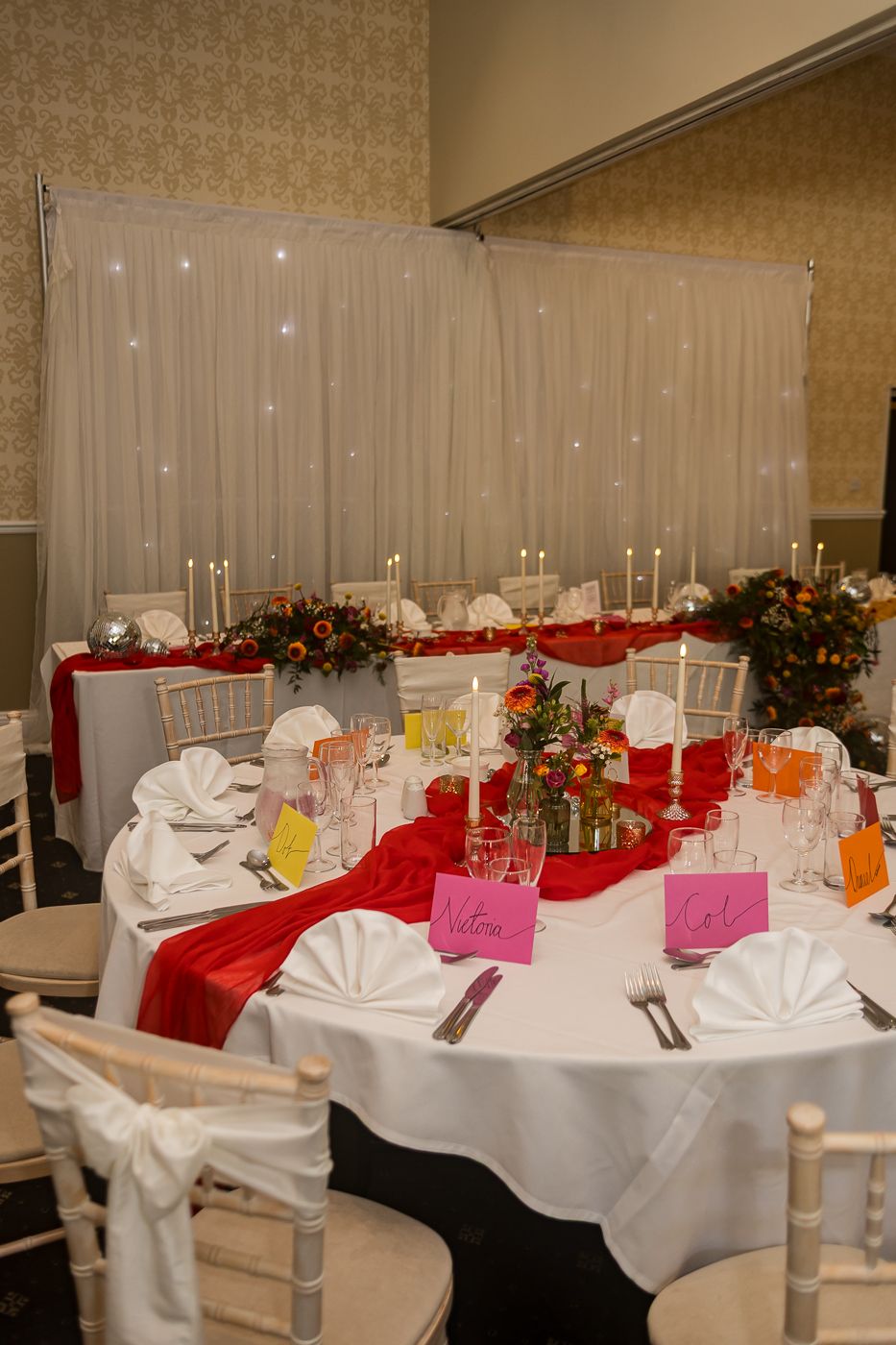 Elegant wedding reception table with red satin runner, white linens, candles, floral centerpiece, and colorful place cards.