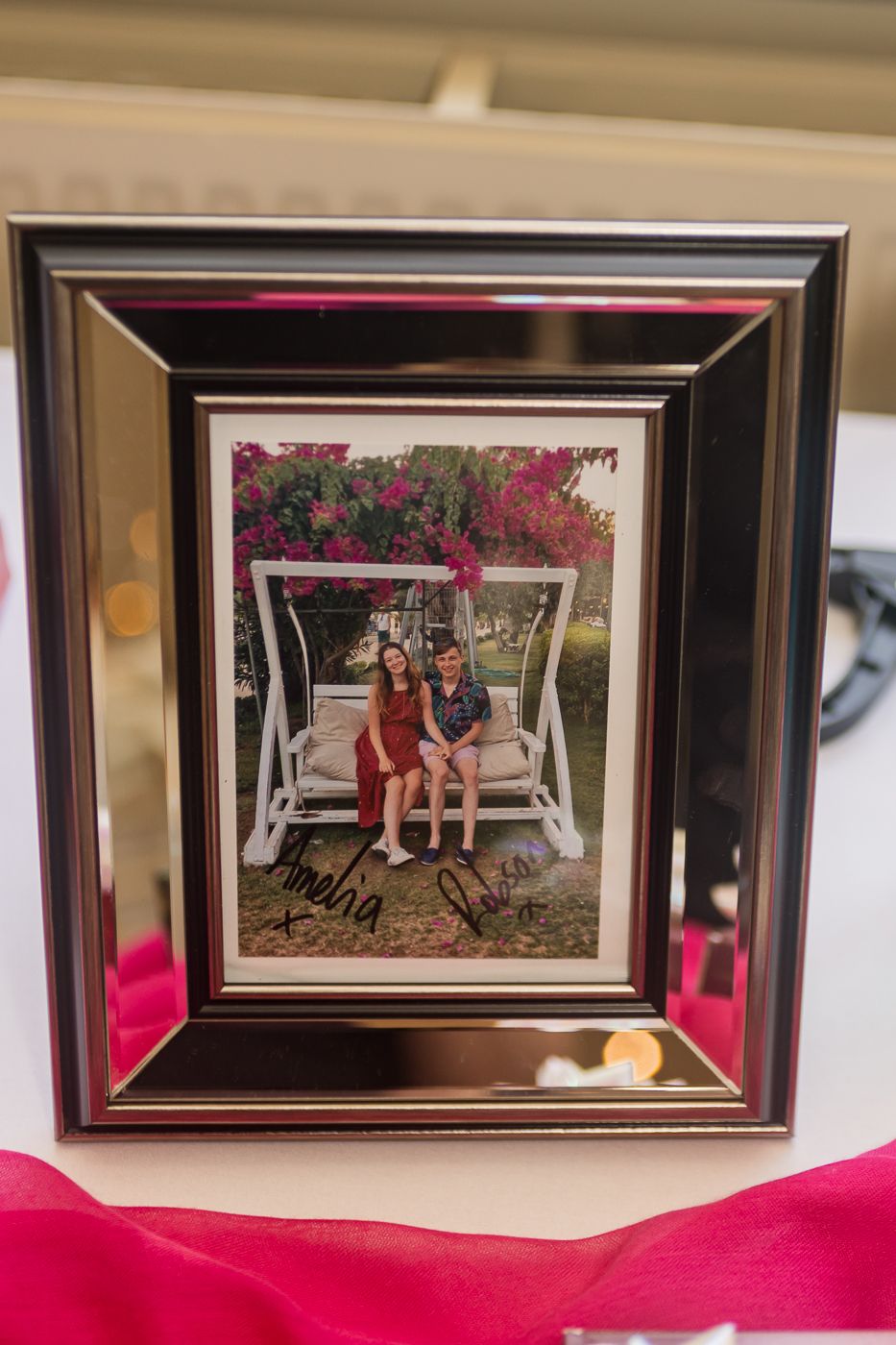 Framed signed photo of a couple sitting on a white swing surrounded by pink flowers displayed on a table.