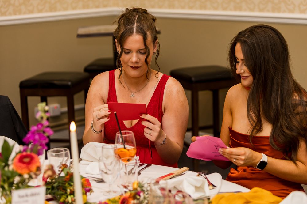 Two women in red dresses sit at a decorated banquet table, examining small cards at a formal event.