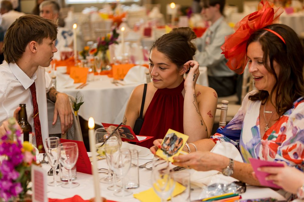 Three women socializing at an elegant event, wearing colorful outfits and fascinators, seated at a decorated banquet table.