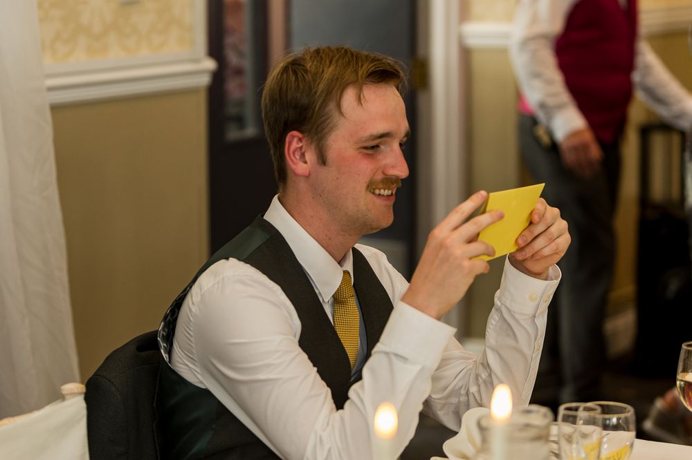 Man in vest and tie smiling while reading a yellow card at a formal dinner event.