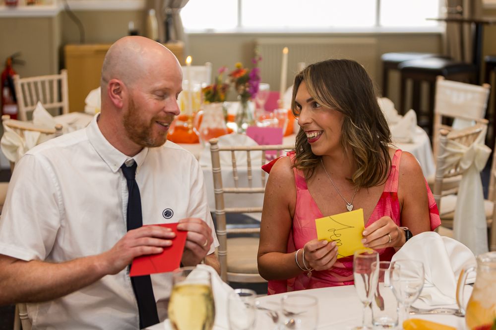 A man and woman smile at each other holding colorful cards at a festive wedding reception dinner table.