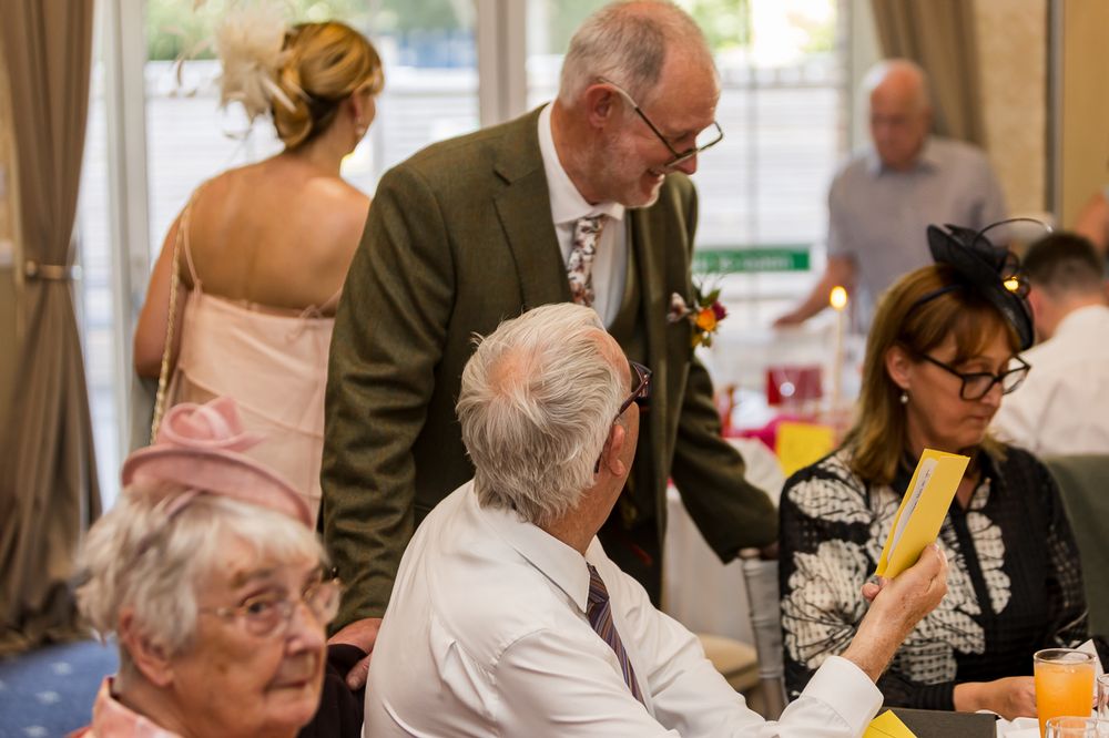 Elderly guests socializing at a formal wedding reception dinner, with women wearing fascinators and men in suits.