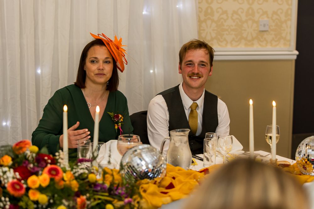 Woman in green dress and orange fascinator sits with mustachioed man in vest at elegant wedding reception table.