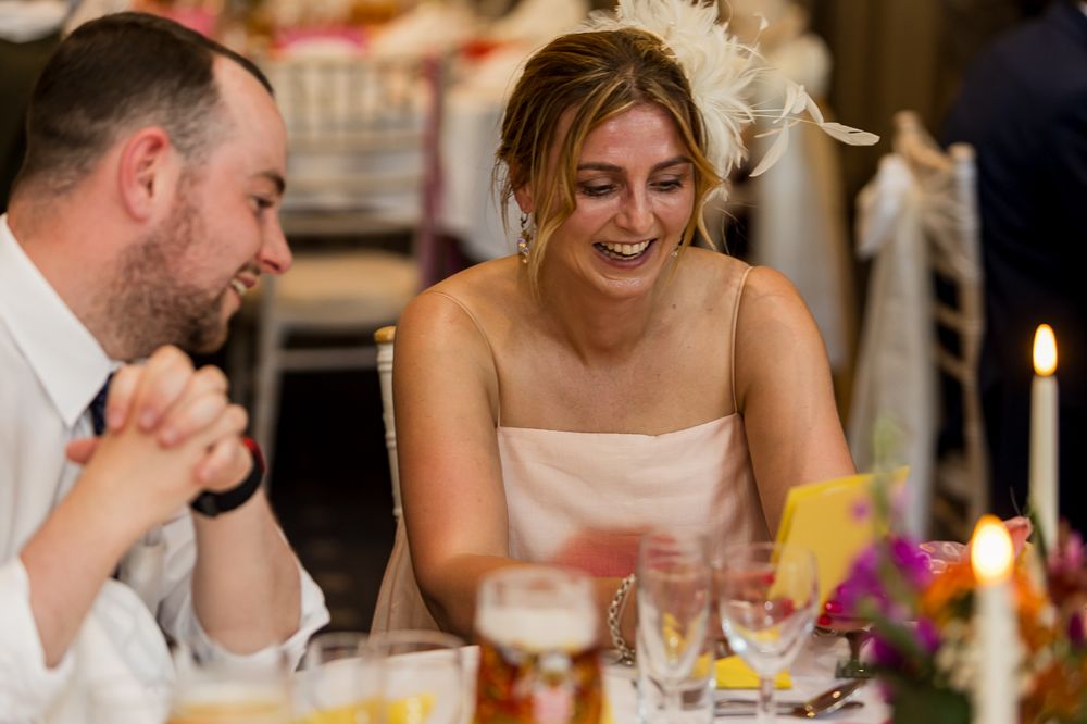 Bride in white dress with feather fascinator laughs joyfully with guest at candlelit wedding reception.