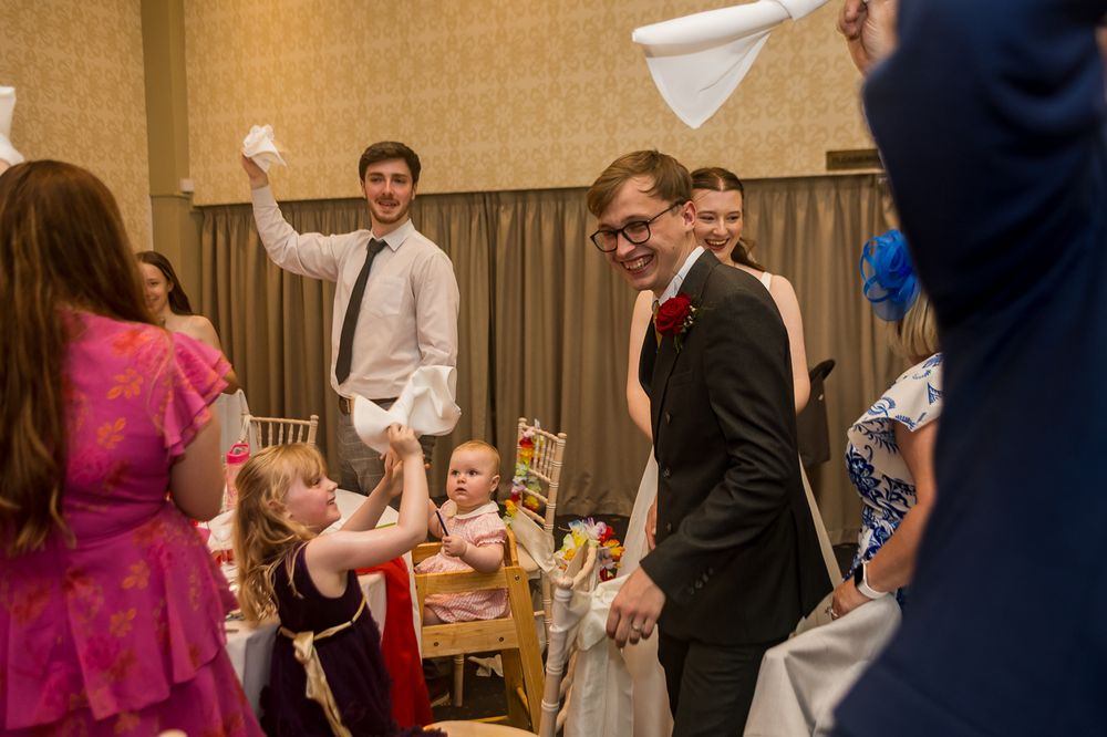 Guests celebrate at a wedding reception, waving white napkins and smiling in a decorated banquet hall.
