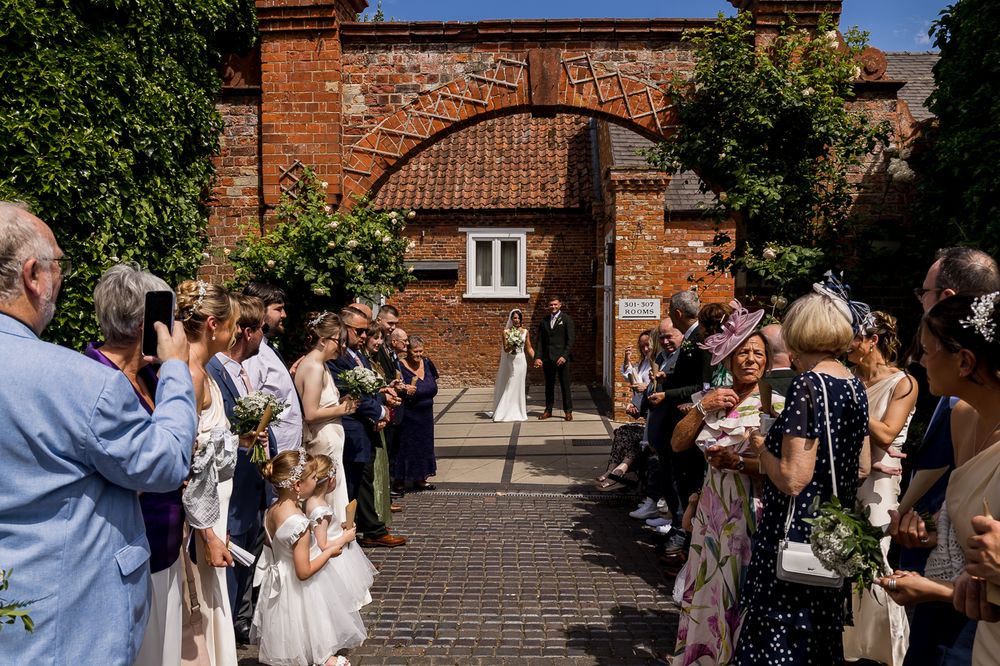 Guests line an outdoor aisle as a bride walks toward the altar at a sunny rustic brick venue wedding ceremony.