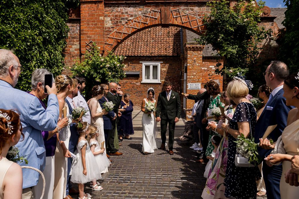 Bride and groom walk through confetti-lined cobblestone path at rustic brick arch wedding venue on sunny day.