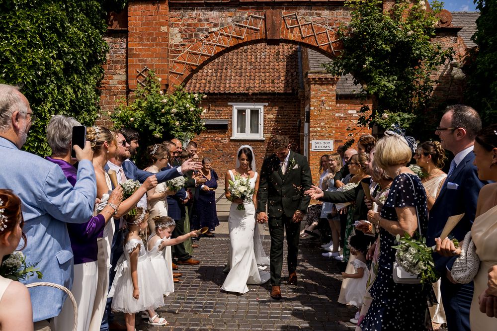 Bride and groom walk through confetti arch at sunny outdoor wedding ceremony with guests cheering.