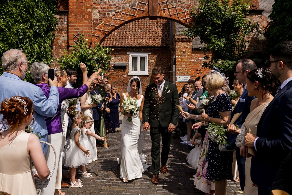 Bride and groom walk through confetti shower surrounded by smiling wedding guests outside a rustic brick venue.