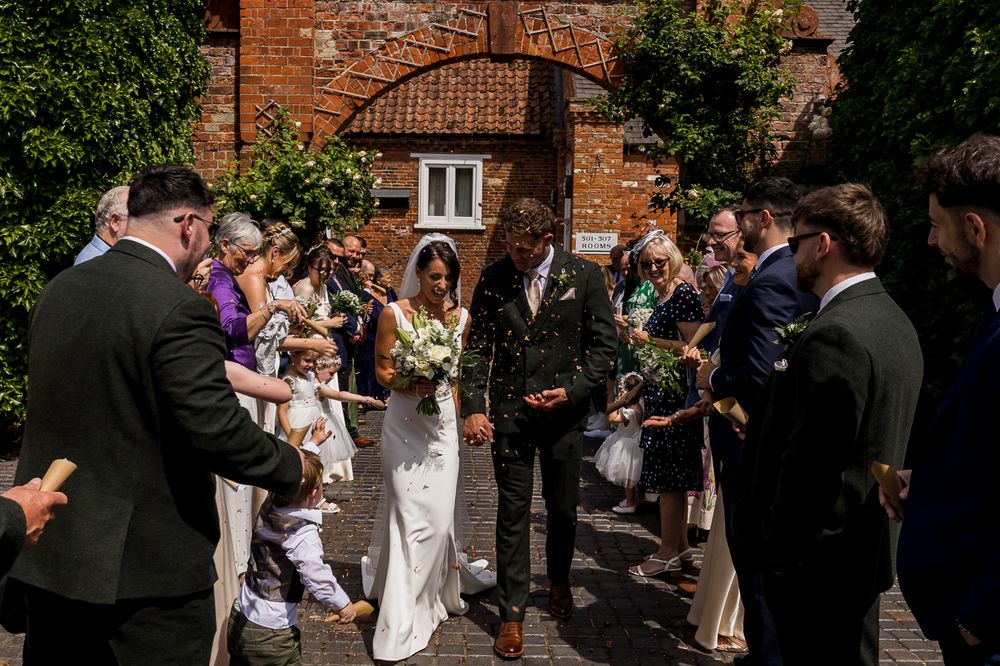 Bride and groom walk through confetti shower with smiling wedding guests outside a rustic brick venue on a sunny day.