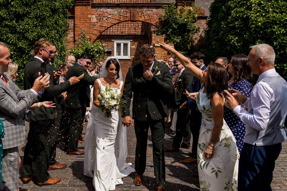 Bride and groom walk through confetti shower surrounded by cheering wedding guests outside a rustic brick venue.