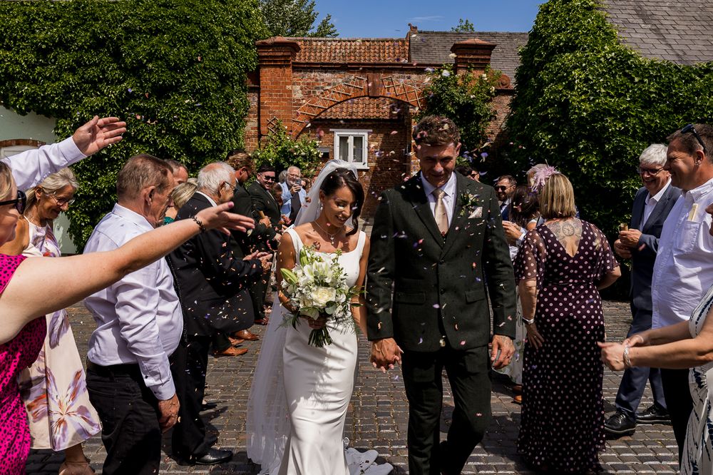 Bride and groom walk through confetti shower surrounded by cheering wedding guests outside a rustic brick venue on a sunny day.