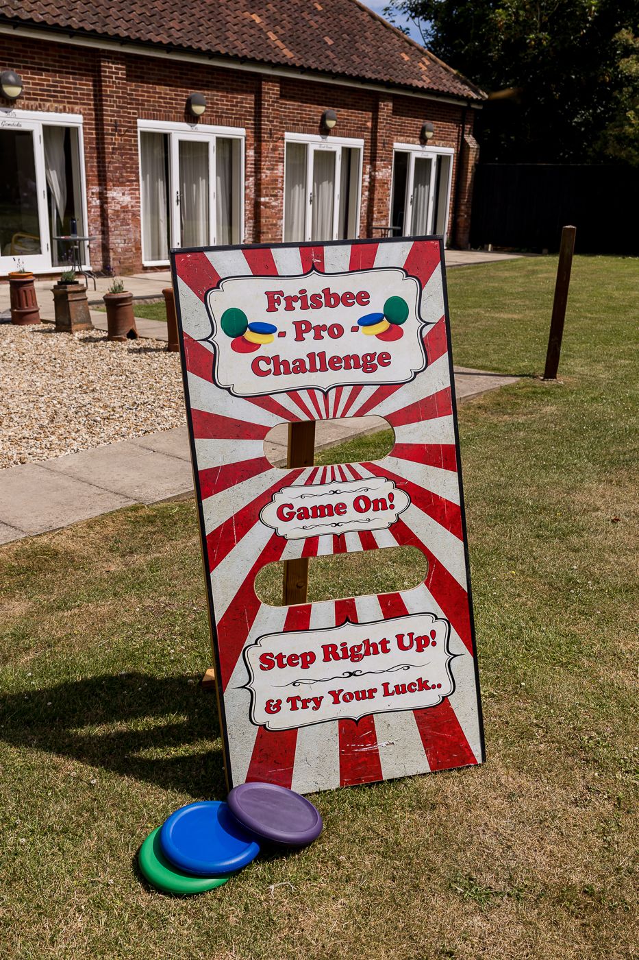 Carnival-style Frisbee Pro Challenge game board with colorful discs on grass outside a brick building.