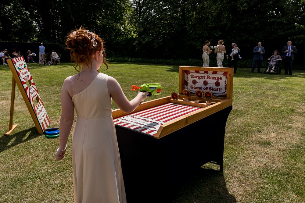 Woman in elegant dress playing carnival target game at outdoor wedding reception on sunny day.