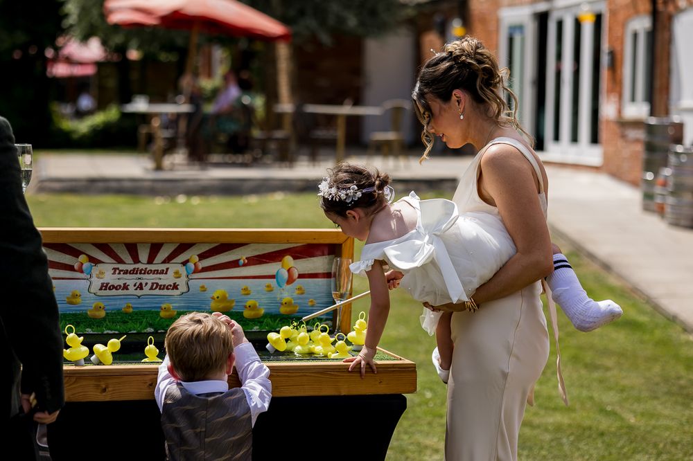 Bride holding baby at duck fishing game with young boy at outdoor wedding reception.