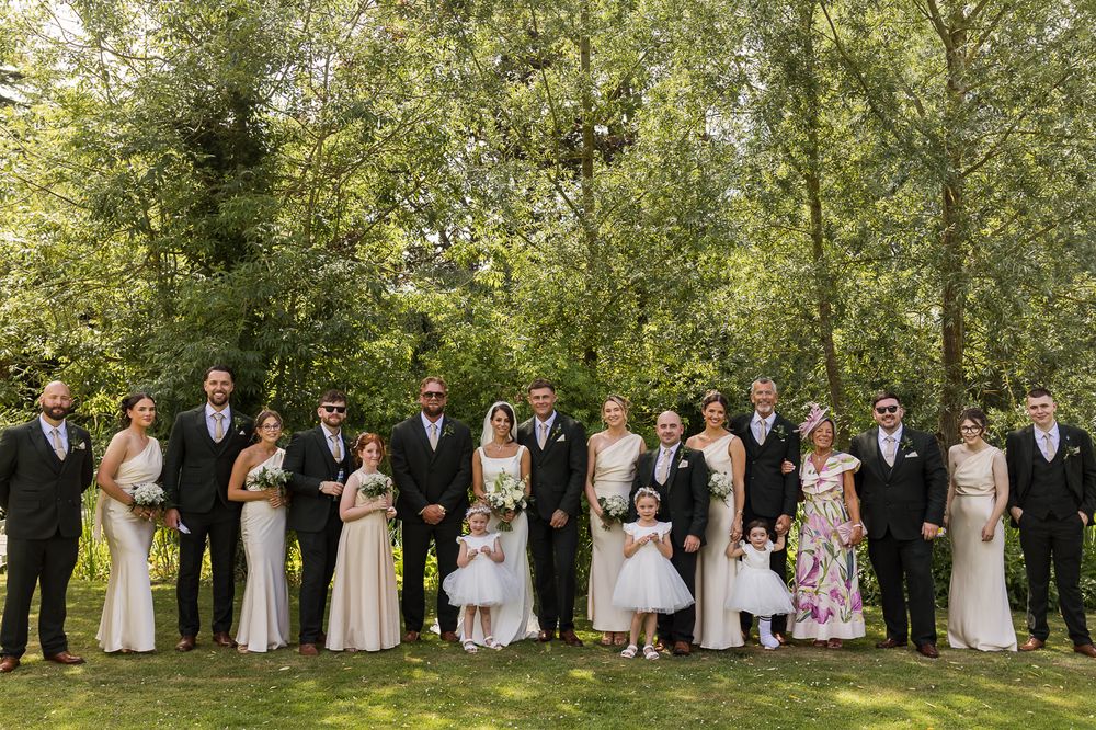 Large wedding party posing outdoors in formal attire, surrounded by lush green trees on a bright summer day.