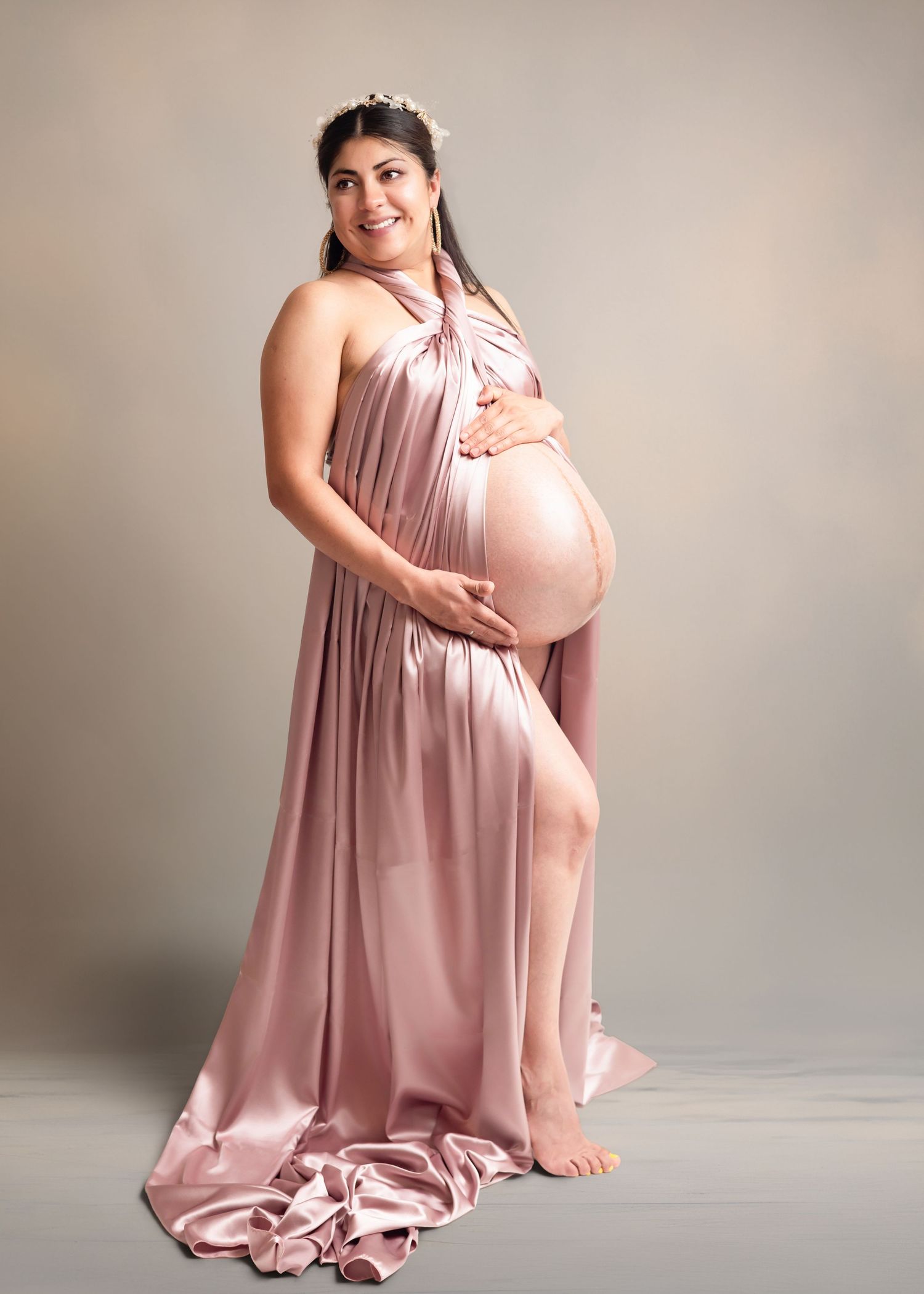Pregnant woman in elegant blush pink satin halter gown cradling baby bump, studio maternity portrait.
