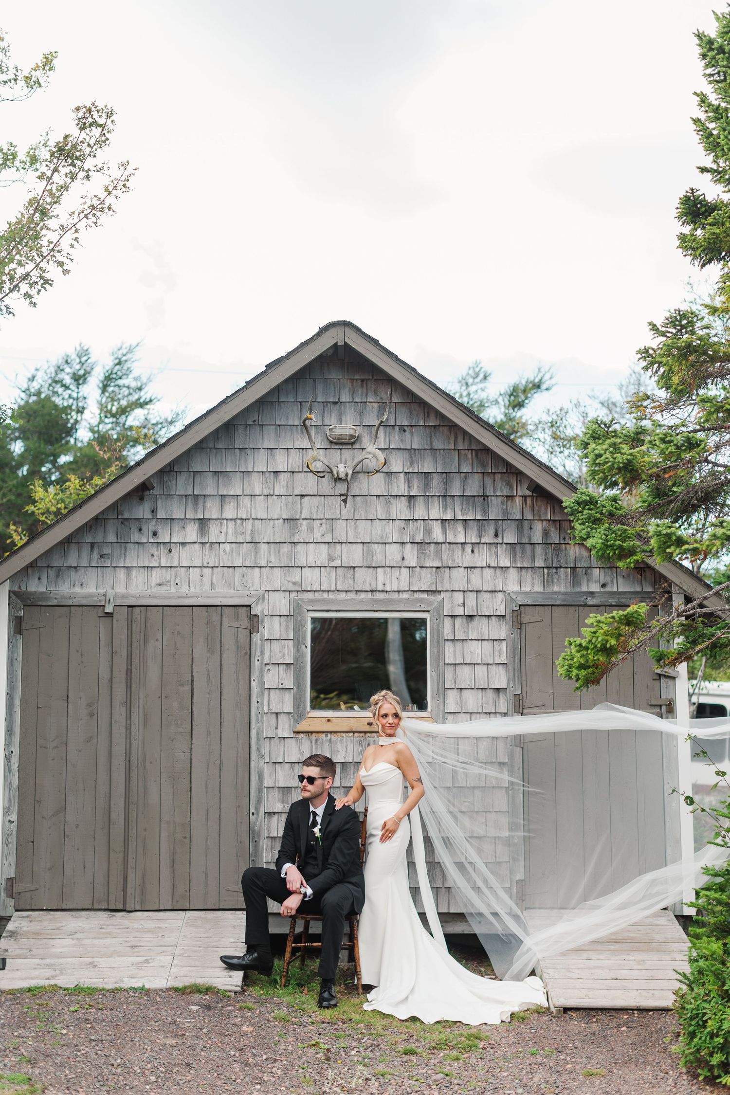 Black and white wedding photo of bride with flowing veil and groom in sunglasses seated outside rustic barn.