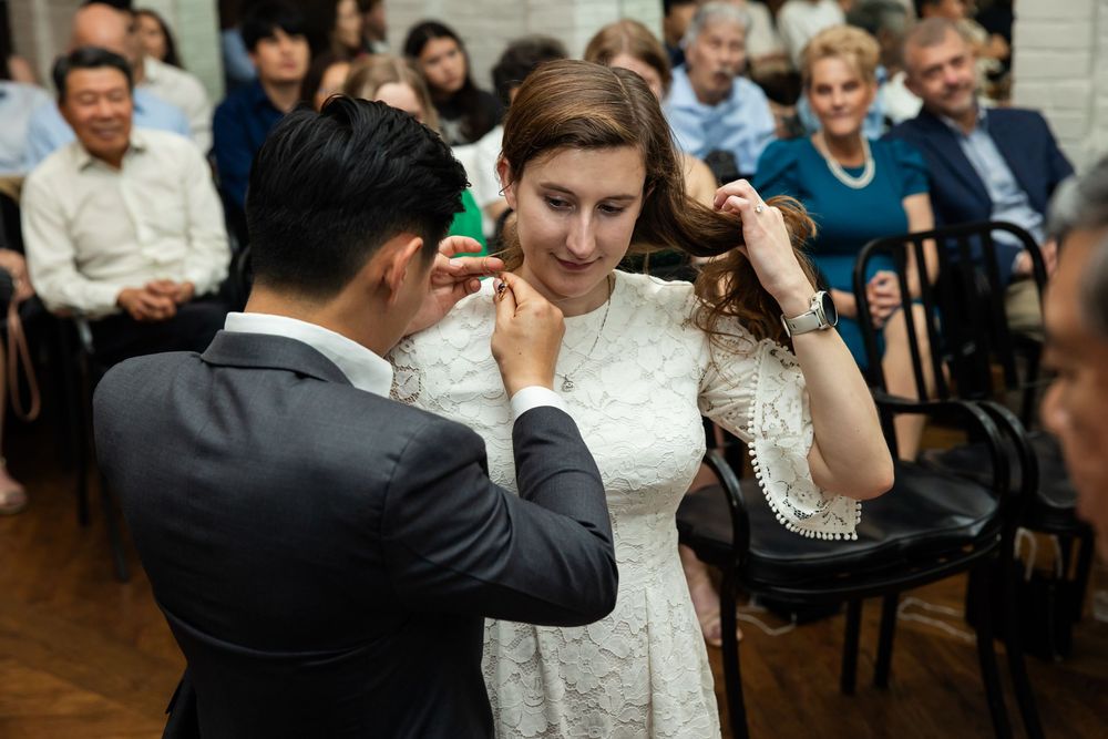 Groom placing earrings on bride in white lace dress during intimate wedding ceremony with seated guests.