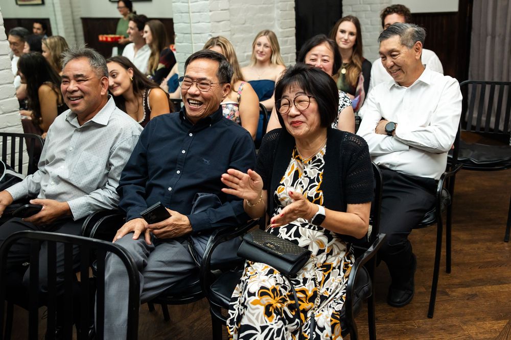 Group of smiling Asian adults laughing and clapping together at an indoor social gathering or event.