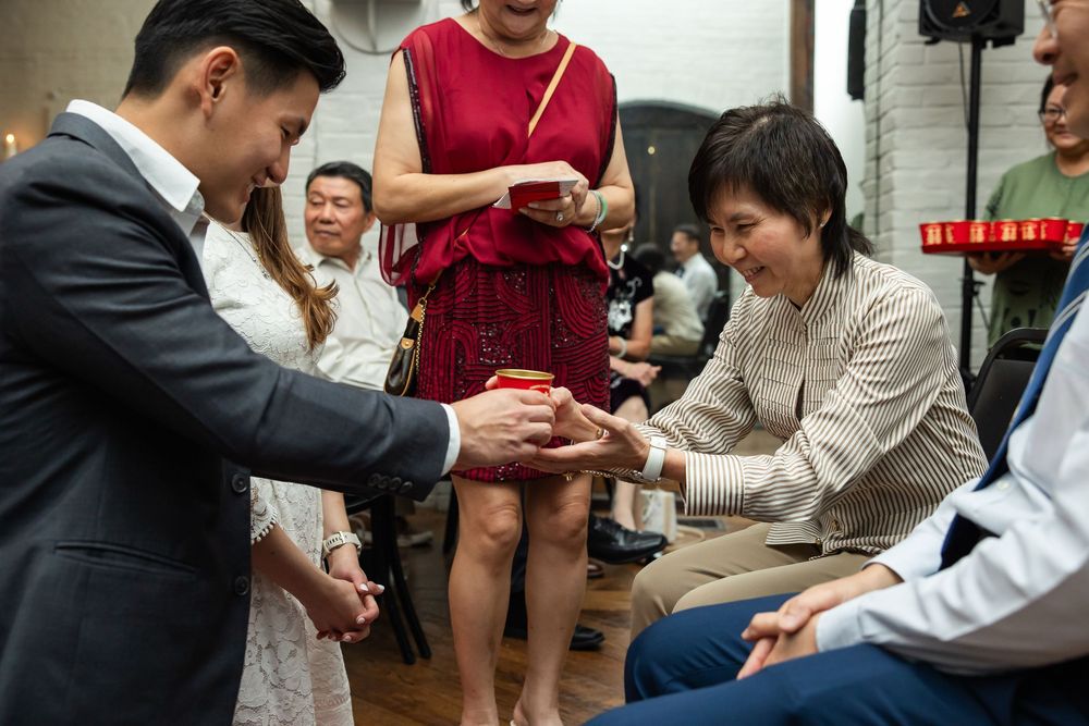 Asian family exchanging gifts during a traditional wedding ceremony indoors, with guests gathered around smiling.