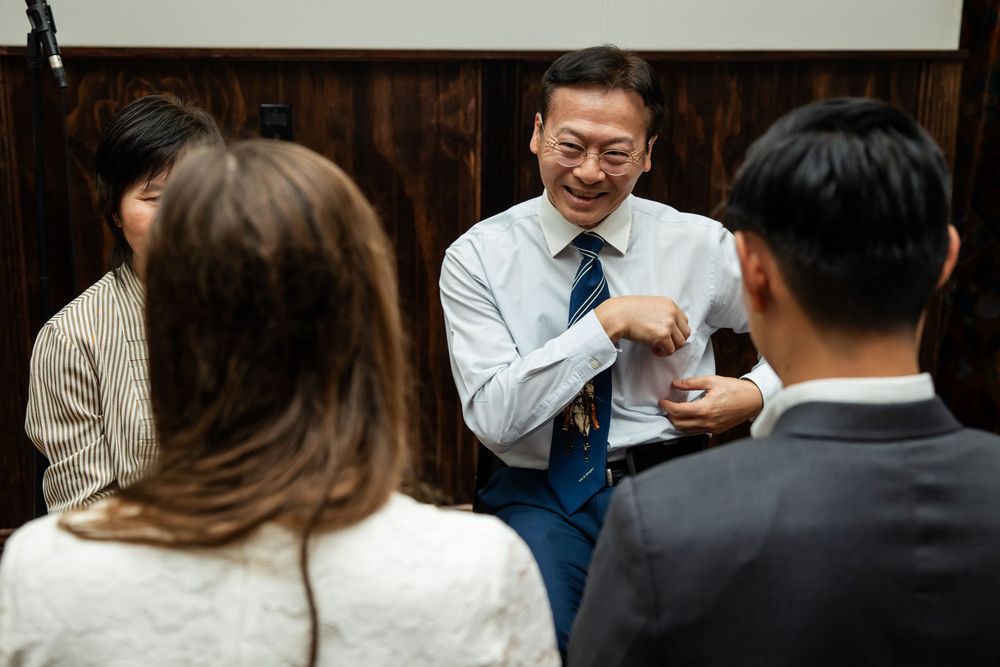 Asian businessman in blue shirt smiling and gesturing during a casual group conversation in a modern office setting.