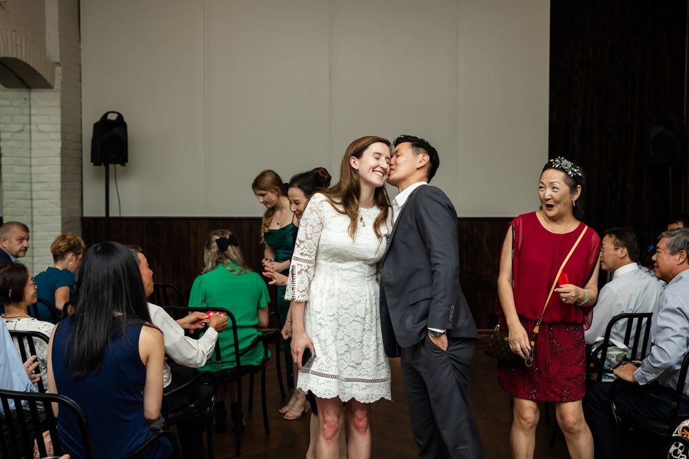 Couple sharing a kiss at a wedding reception with guests socializing in the background.