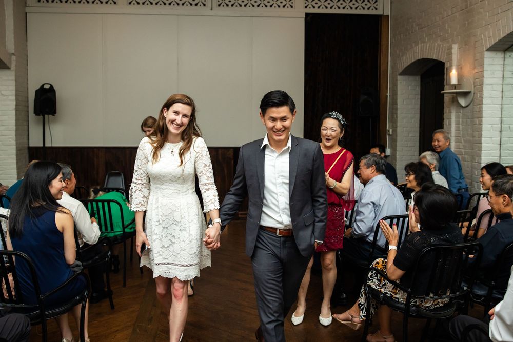 Couple holding hands walking together at a wedding reception venue with guests seated in the background.