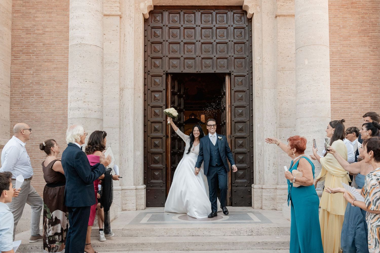 Bride and groom exit a historic church through ornate wooden doors as smiling wedding guests cheer and applaud on the stone steps.