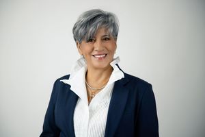 Smiling woman with short gray hair wearing a navy blazer over a white ruffled collar blouse and a delicate necklace against a light backgrou